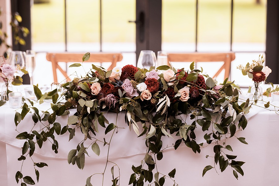 a beautiful floral arrangement with pale pink and dark red flowers and trailing greenery sits on the top table at a wedding at Middleton Lodge in North Yorkshire.