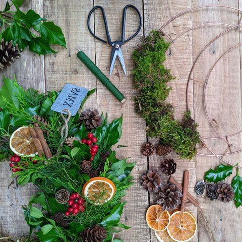 A flat lay of wreath-making materials on a wooden surface, including greenery, pinecones, dried orange slices, moss, scissors, and a partially assembled wreath.