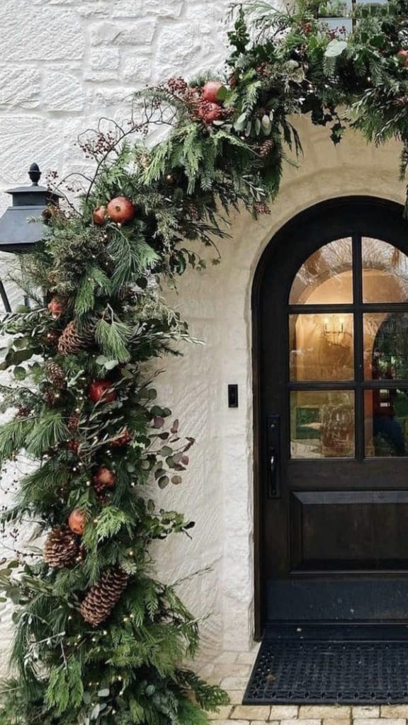 Evergreen garland with pinecones and red ornaments decorates the arched doorway of a white stone house with a black door and outdoor lantern.