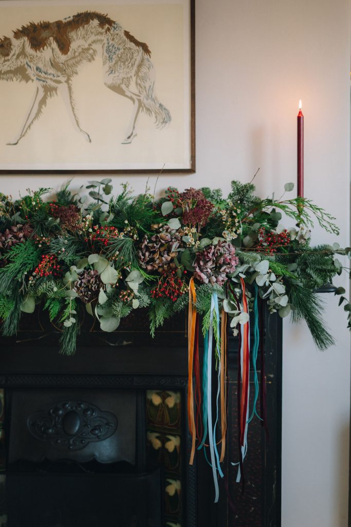 Festive garland with pine, berries, and ribbons decorates a mantel. A single red candle is lit, and animal artwork hangs above.