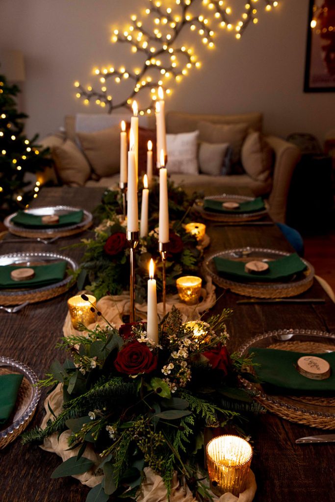 A dining table set for six with green napkins, glass plates, gold candles, and floral centerpieces, with string lights and a sofa in the background.