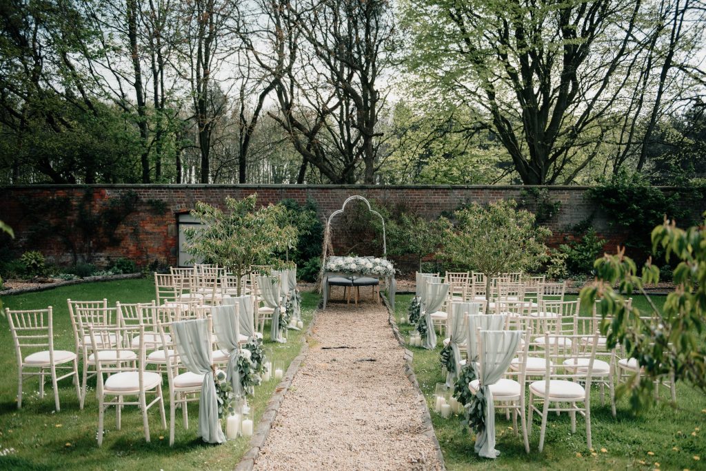 Outdoor wedding ceremony setup with rows of white chairs, a gravel aisle, floral decorations, and an arch with greenery, set in a garden with trees and a brick wall in the background.