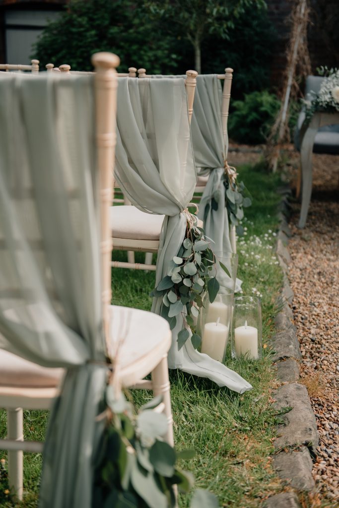 Rows of wooden chairs draped with light green fabric and eucalyptus leaves, with glass candles on the grass, set up for a beautiful outdoor ceremony at Shotton Grange in Northumberland.