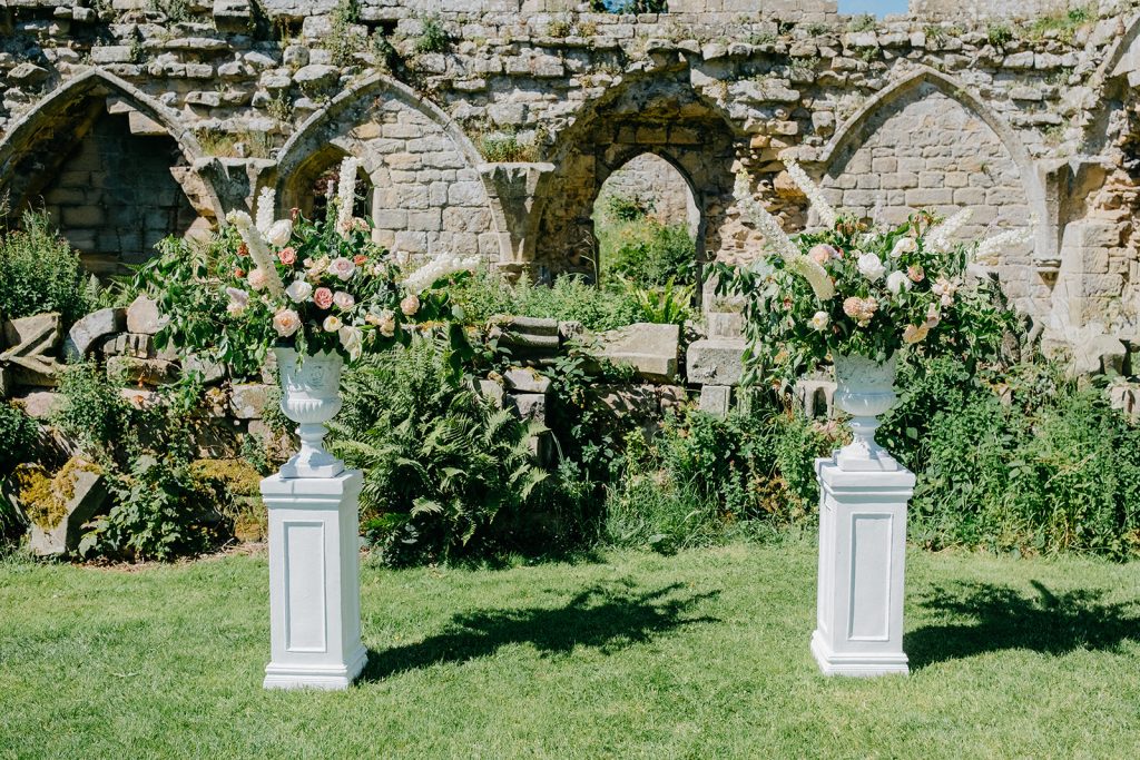 Two white pedestals with outdoor wedding ceremony flowers stand on a lawn in front of the old stone ruins and arched openings of Jervaulx Abbey, surrounded by lush greenery.