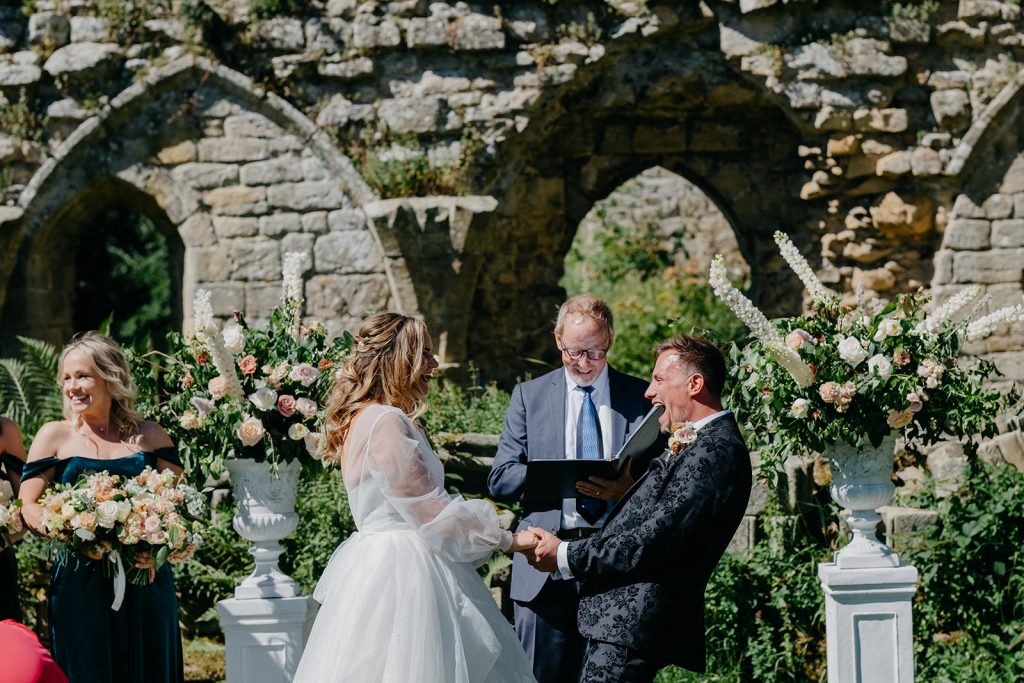 A couple stands holding hands and smiling during an outdoor wedding ceremony at Jervaulx Abbey, with an officiant reading and guests nearby, framed by a stone wall and elegant floral arrangements.