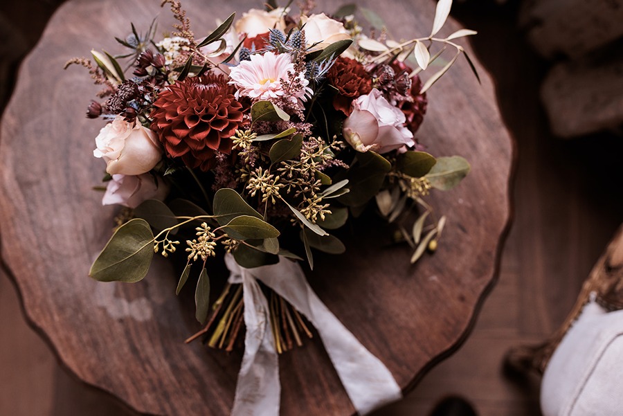 An elegant Autumn bridal bouquet of red, pink, and white flowers with green leaves and a white ribbon, beautifully arranged on a round wooden table at Middleton Lodge.