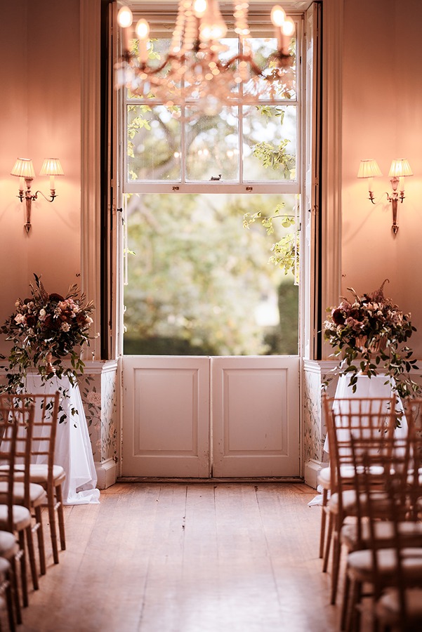 Elegant indoor Main House wedding ceremony setup at Middleton Lodge, featuring rows of wooden chairs, beautiful floral arrangements, and large windows with natural light filtering in.