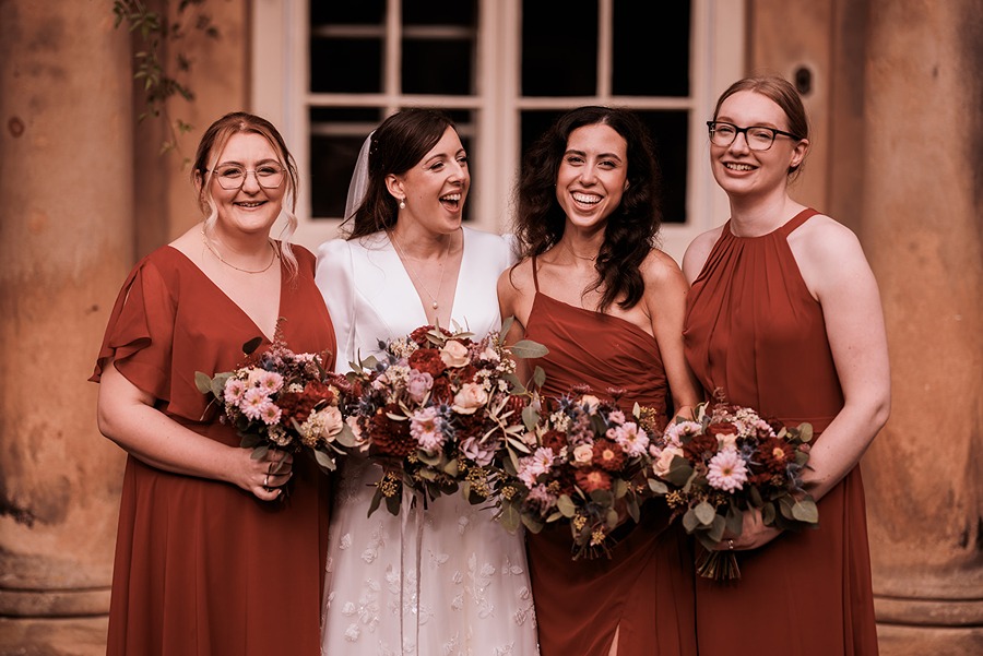 Four women pose in front of Middleton Lodge; one in a white wedding dress and the other three in matching reddish dresses, all holding floral bouquets and smiling after a beautiful Main House wedding ceremony.
