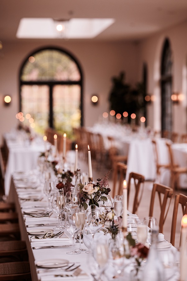 Long banquet tables at Middleton Lodge are set with white tablecloths, elegant glassware, exquisite wedding flowers, and lit taper candles in a beautifully decorated room with arched windows.