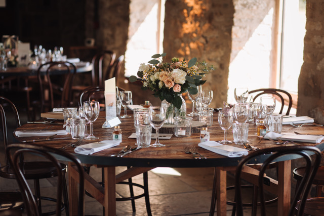 A rustic wooden table in the Healey Barn dining room is set for a formal meal with glassware, plates, cutlery, white napkins, and elegant wedding flowers arranged by a skilled florist, in a softly lit room with stone walls and large windows.
