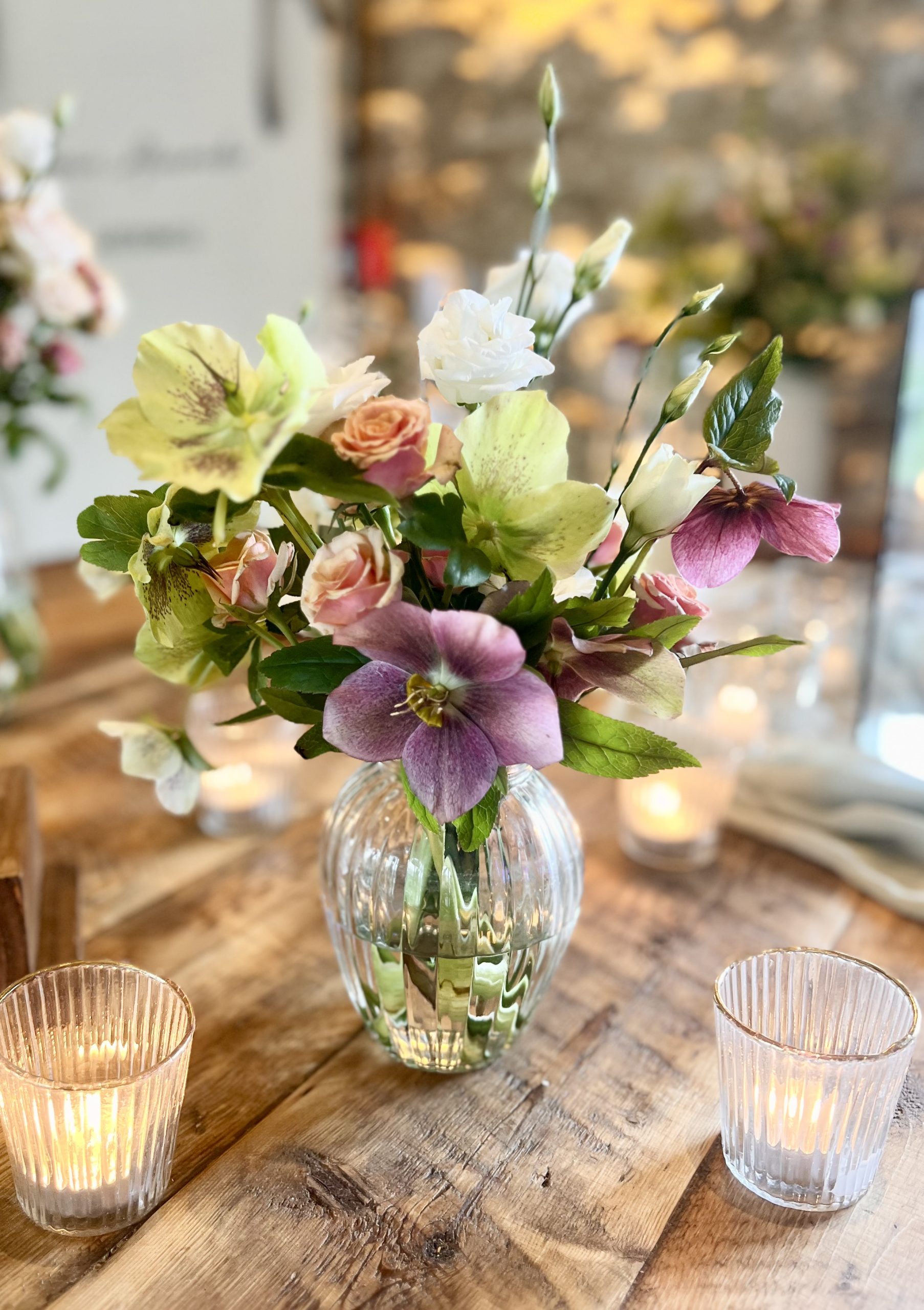 A glass vase with a mixed bouquet of wedding flowers, arranged by a skilled florist, sits on a wooden table in the Healey Barn dining room between two lit candles in ribbed glass holders.