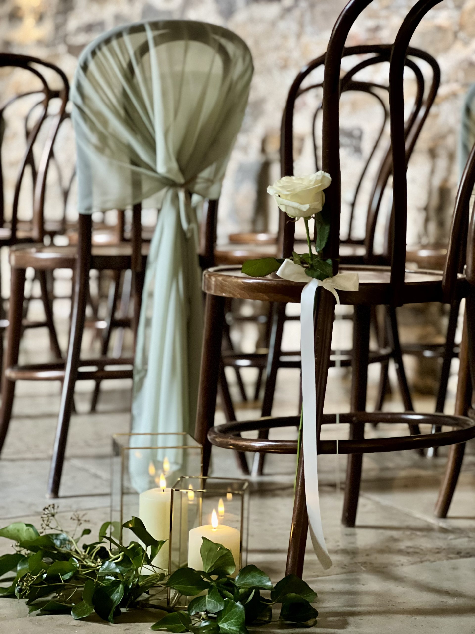 Rows of wooden chairs in classic aisle style, one draped with light fabric, with a white rose and greenery tied to another—perfect for an intimate wedding ceremony at Healey Barn. Lit candles and leaves decorate the floor nearby.