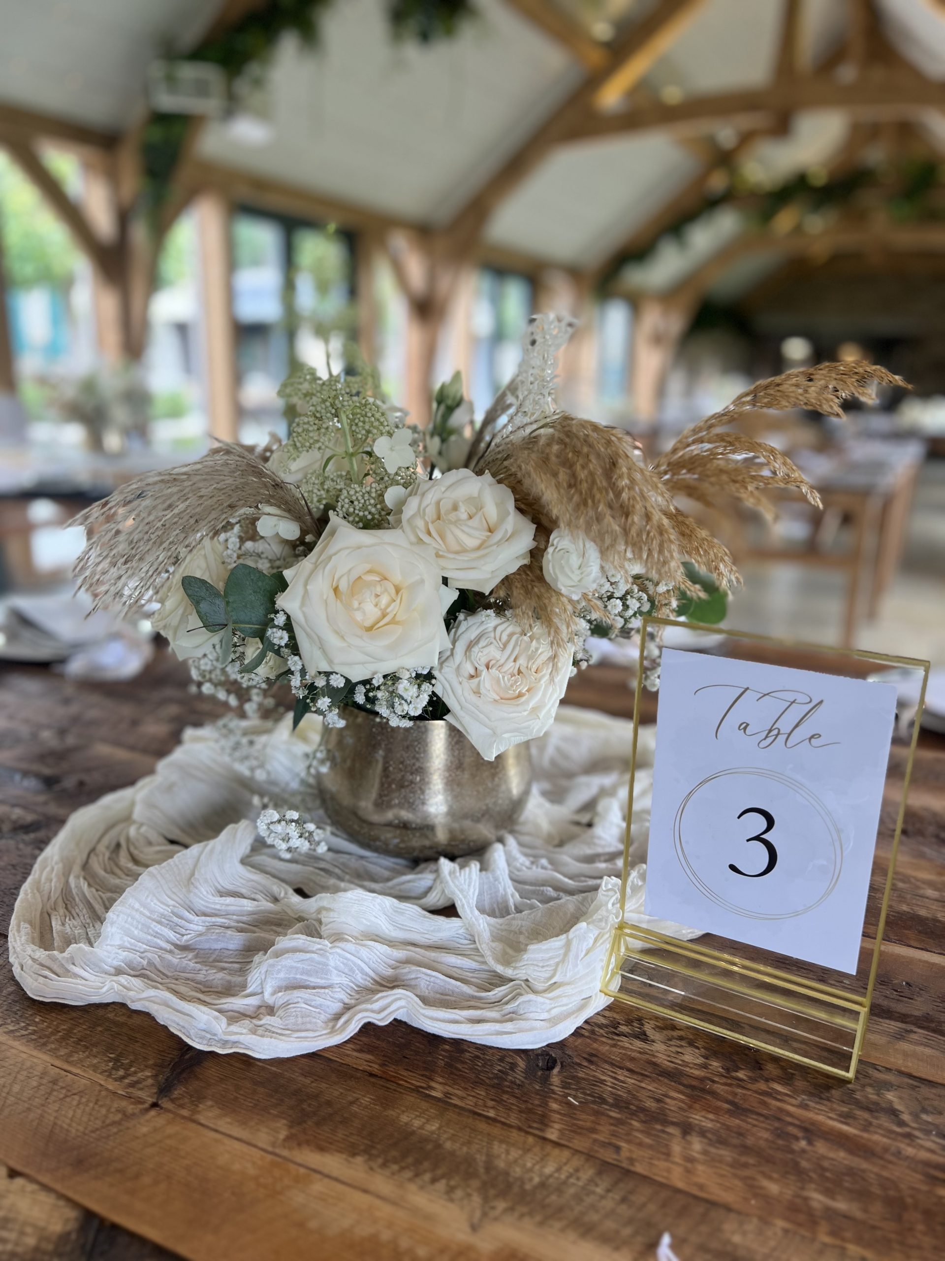 A wedding centrepiece of white roses, greenery, and dried grasses in a metallic vase sits beside a 