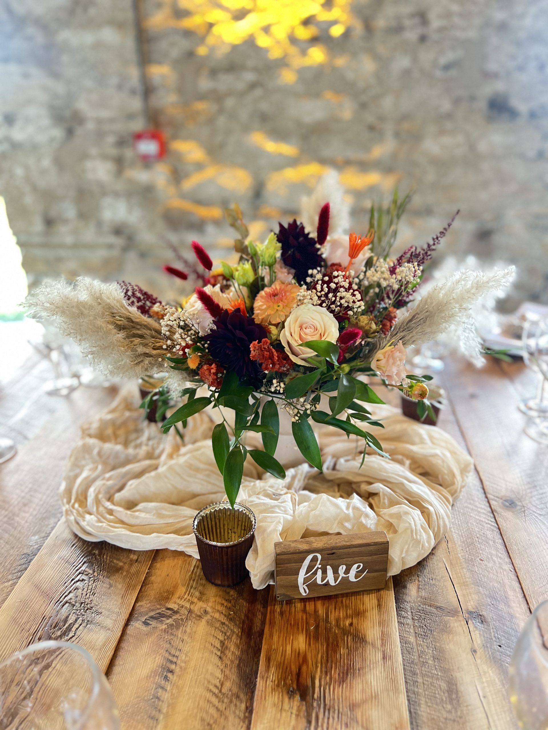 A floral centerpiece with mixed wedding flowers sits on a rustic wooden table in the Healey Barn dining room, next to a candle and a wooden sign displaying the word 
