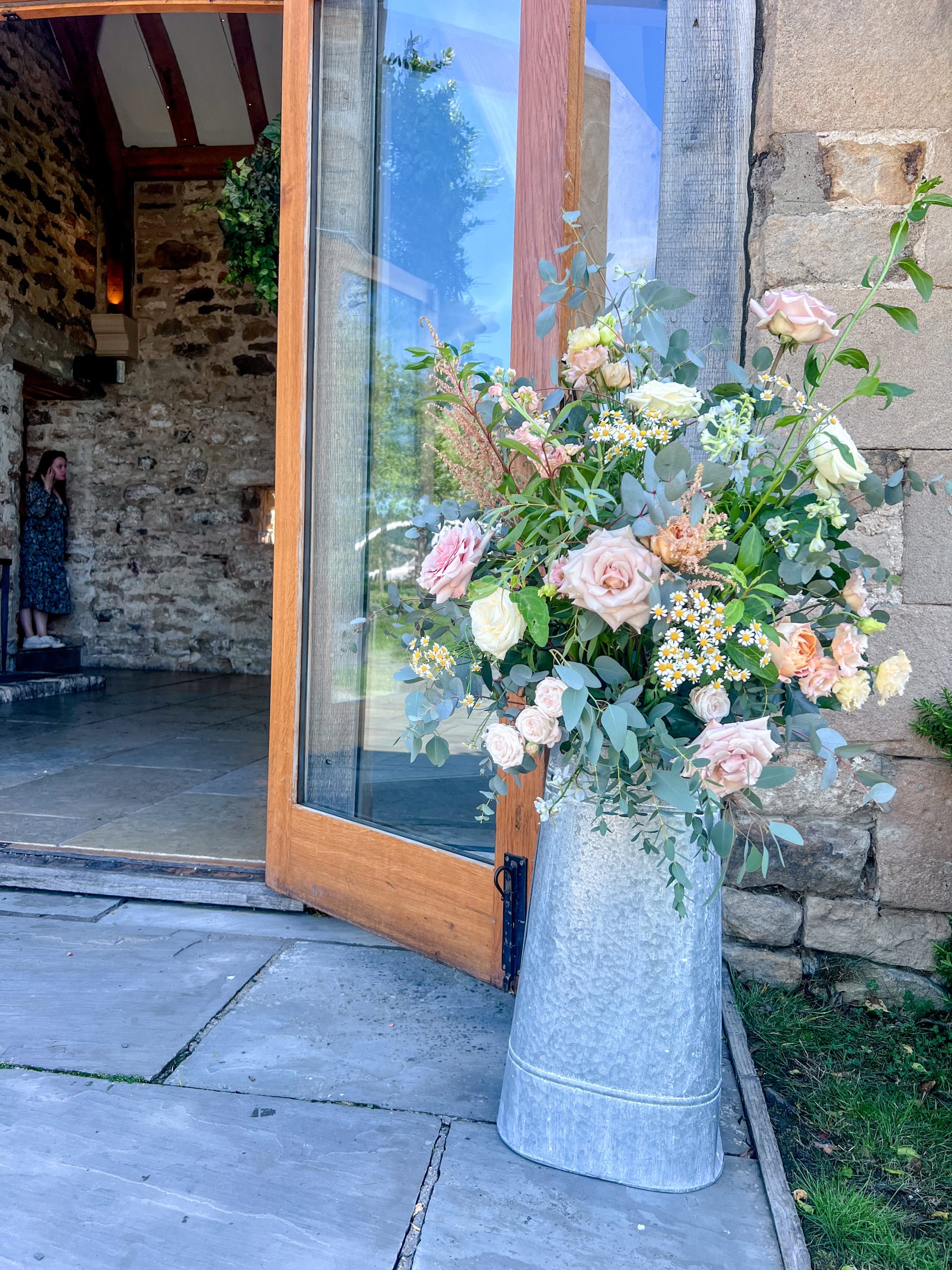 A large floral milk churn filled with pink and white flowers stands on stone paving beside an open wooden door leading into Healey Barn, perfect for a romantic wedding ceremony in a rustic stone building.