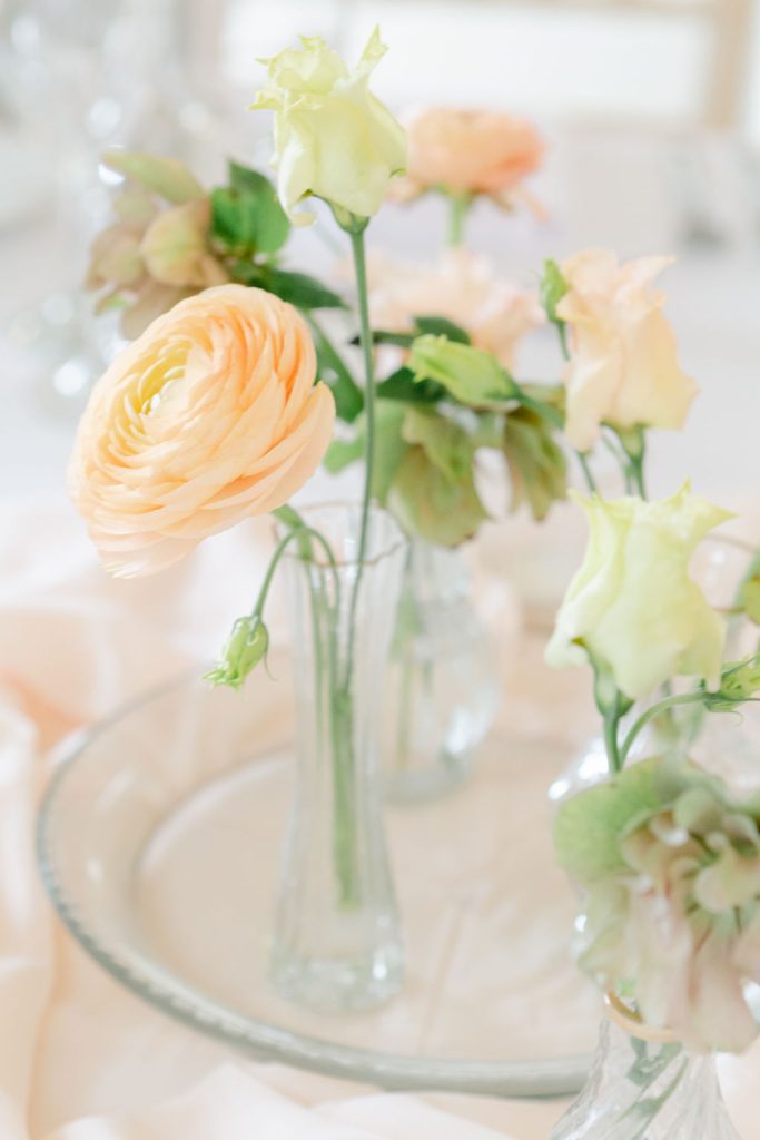 A clear glass vase holds pale peach and light green spring wedding flowers, creating an elegant Northumberland wedding centrepiece on a glass plate with a soft, light background.