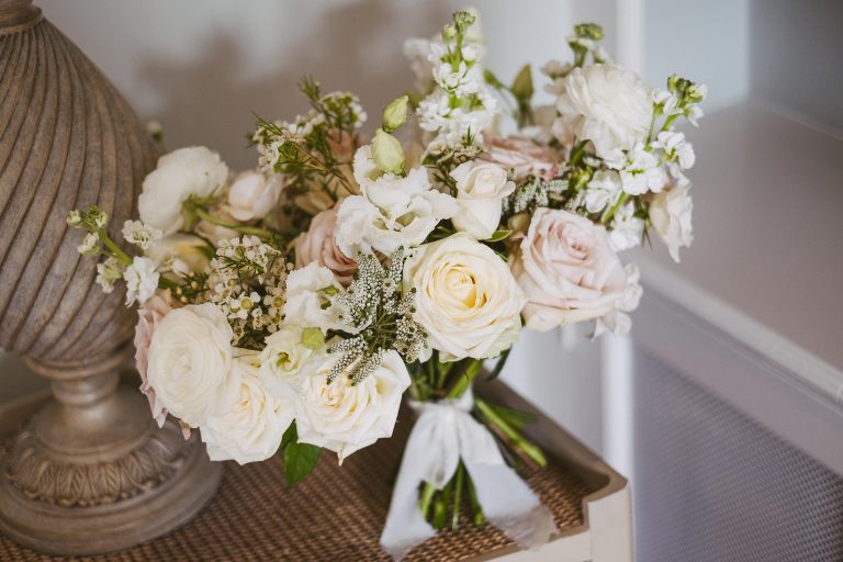 A bridal bouquet of white and pale pink roses with greenery, tied with a white ribbon, rests on a woven surface next to a decorative lamp—perfect inspiration for wedding flowers at Wynyard Hall.