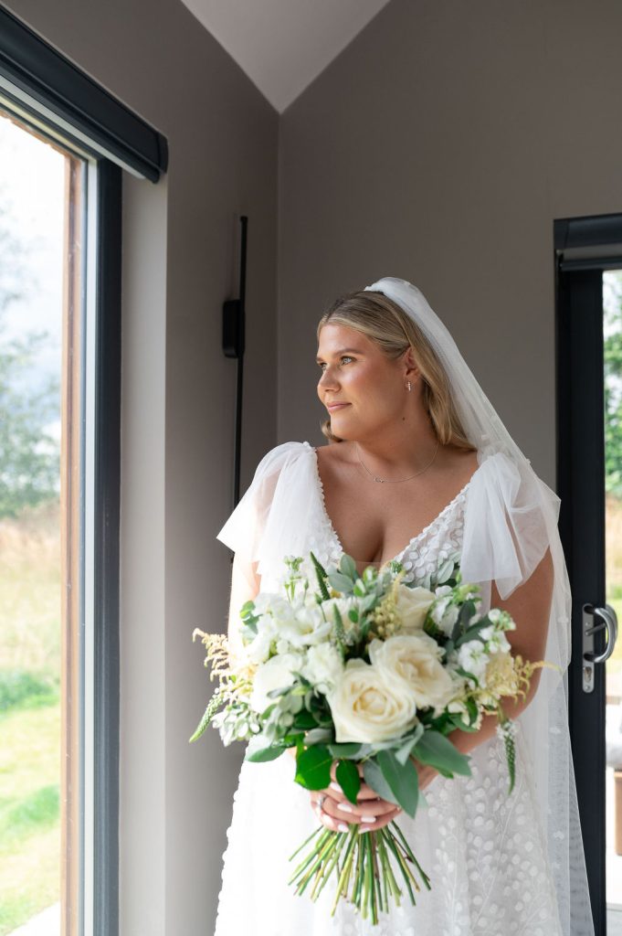 A bride in a white dress and veil holds a white bridal bouquet of roses and greenery, standing indoors near a window at Healey Barn and looking outside.