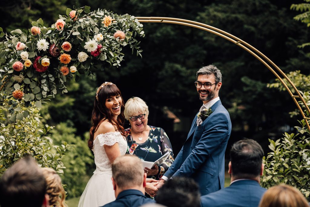 A bride and groom stand under a floral arch, holding hands and smiling, with an officiant reading from a book in front of guests at an outdoor wedding ceremony.