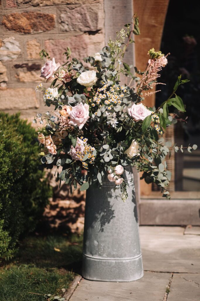 A large metal vase, arranged by a skilled florist, holds pink roses, small white flowers, eucalyptus, and greenery on a stone patio near a brick wall—perfect for an elegant Healey Barn wedding ceremony.