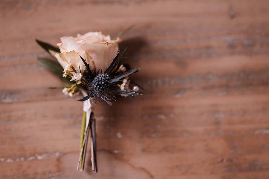 A groom’s buttonhole with pale pink roses, white buds, and a blue thistle, resting on a rustic wooden surface—perfect for wedding flowers at Middleton Lodge.