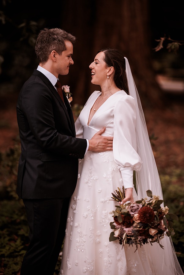 A bride in a white dress and veil holds wedding flowers and smiles at a groom in a black suit and tie; they stand close together outdoors at Middleton Lodge in North Yorkshire.
