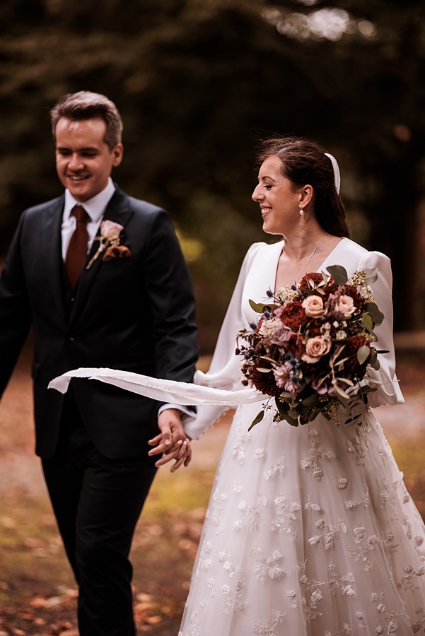 A bride in a white dress holding a beautiful bridal bouquet walks outdoors with a groom in a dark suit at Middleton Lodge; both are smiling.