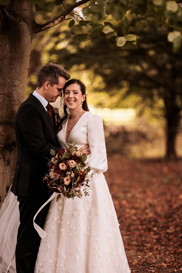 A bride in a white dress holding a stunning bridal bouquet stands beside a groom in a black suit under a tree at Middleton Lodge, with autumn leaves scattered on the ground.