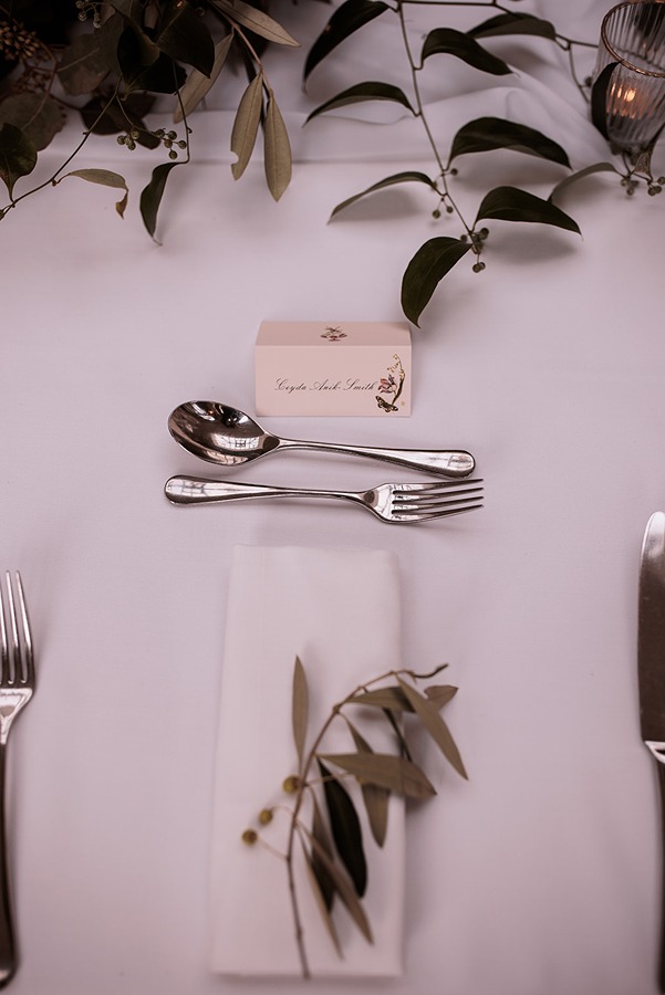 Place setting with spoon, knife, and fork on a white tablecloth, a serviette adorned with wedding flowers and greenery, plus a name card—perfect for an elegant celebration at Middleton Lodge in North Yorkshire.