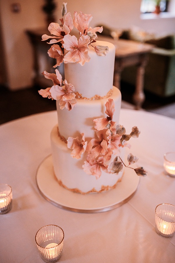 Three-tier white Middleton Lodge wedding cake adorned with delicate pink flowers, placed on a round table surrounded by four small lit candles.