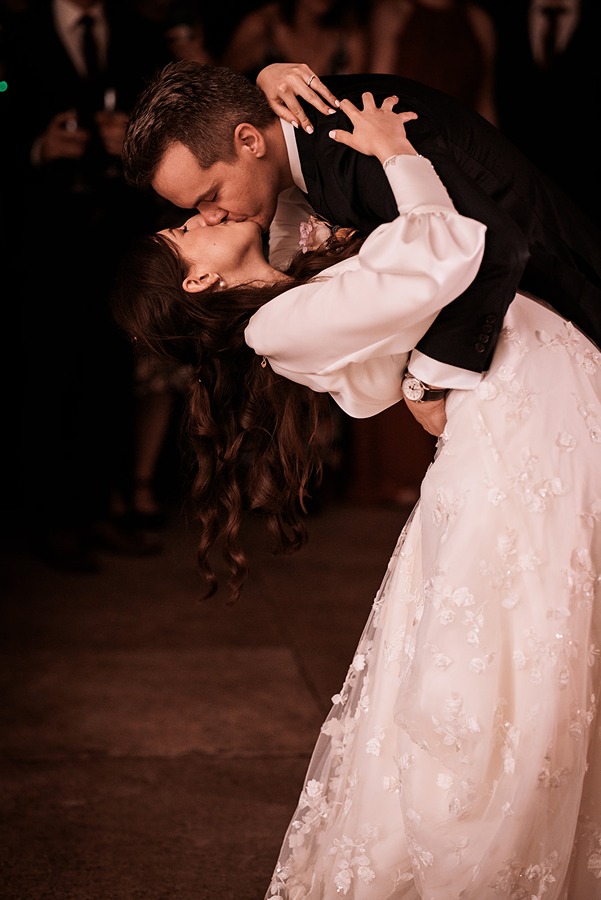 A groom in a suit dips and kisses his bride, who is wearing a white floral wedding dress, during a dance at their wedding reception.