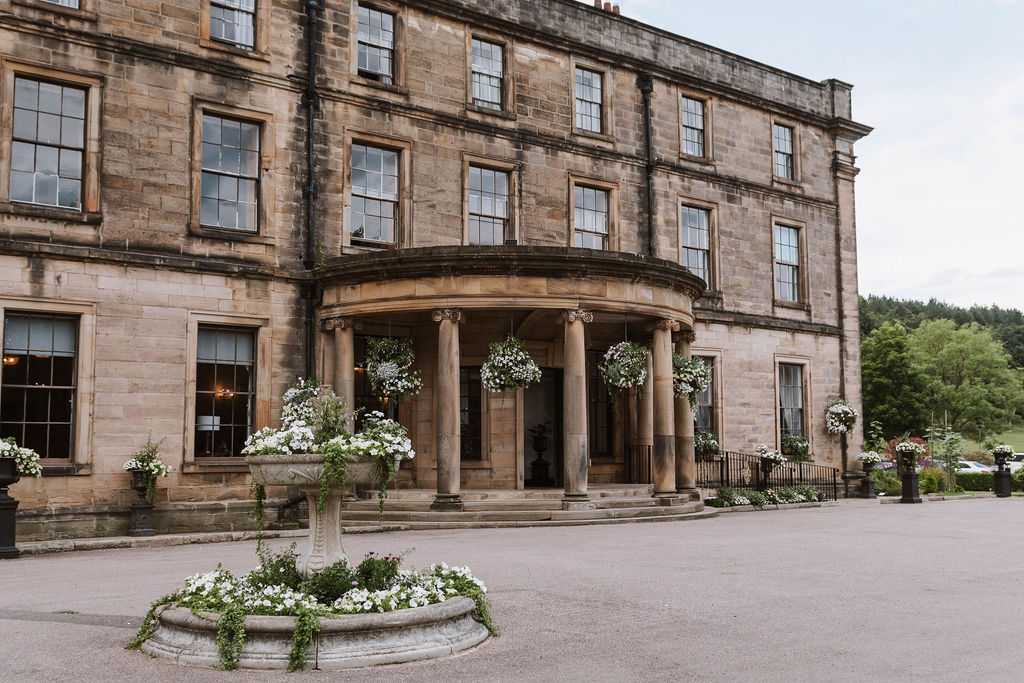 A large stone building with columns at the entrance, tall windows, hanging flower baskets, and a circular flower bed in front.