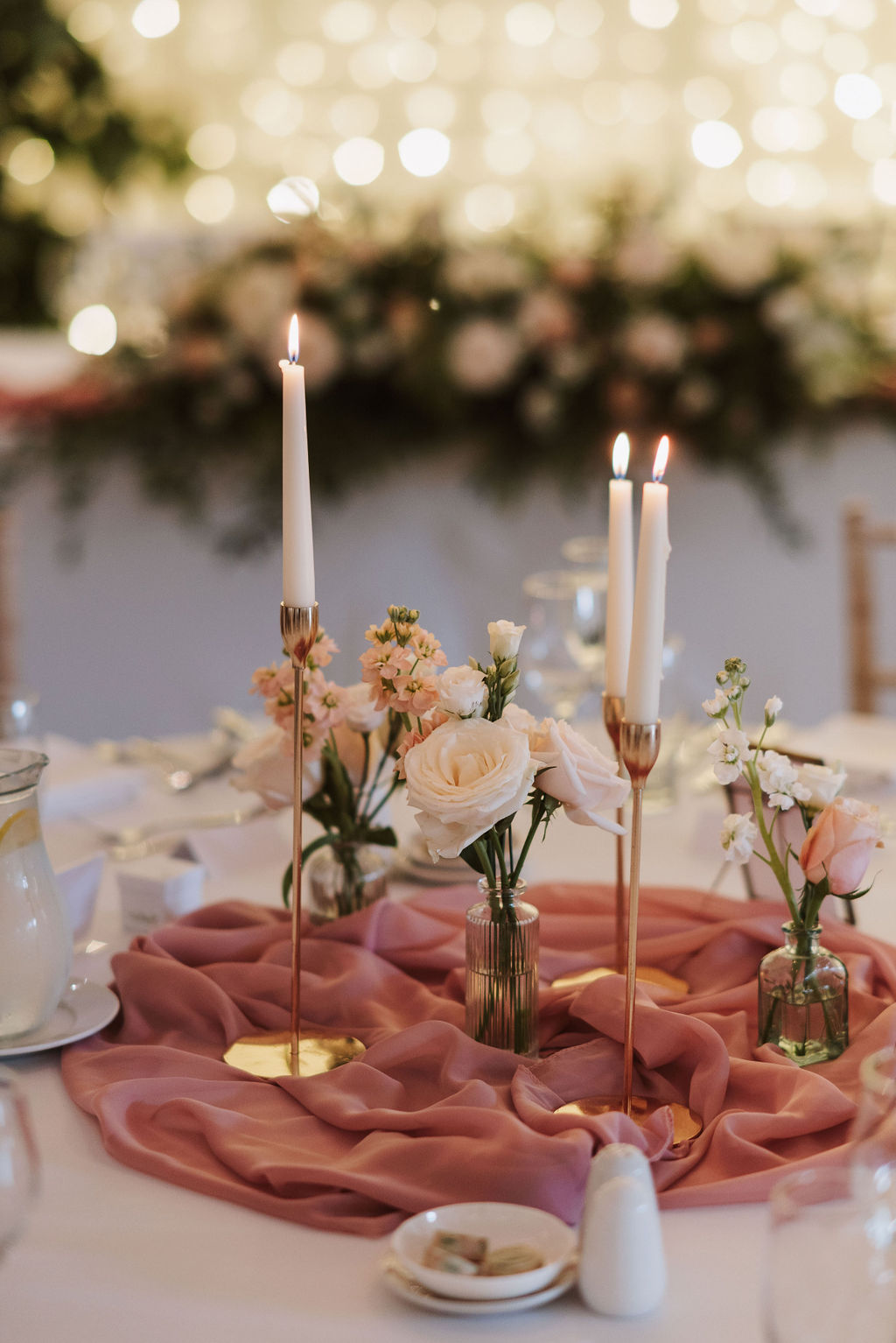 A round table centrepiece with white and blush flowers in glass vases, gold candle holders with tall lit candles, and a mauve sheer cloth, set for an event or celebration.