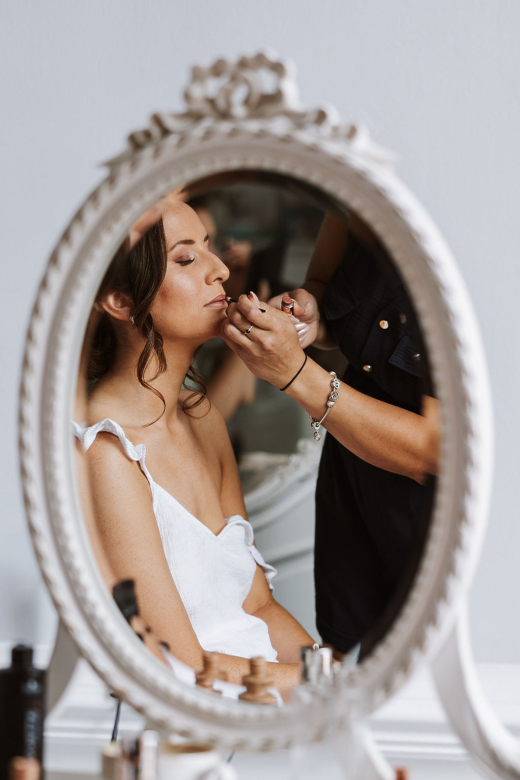 A woman in a white dress sits with her eyes closed whilst another person applies make-up to her lips, as seen through an ornate oval mirror.