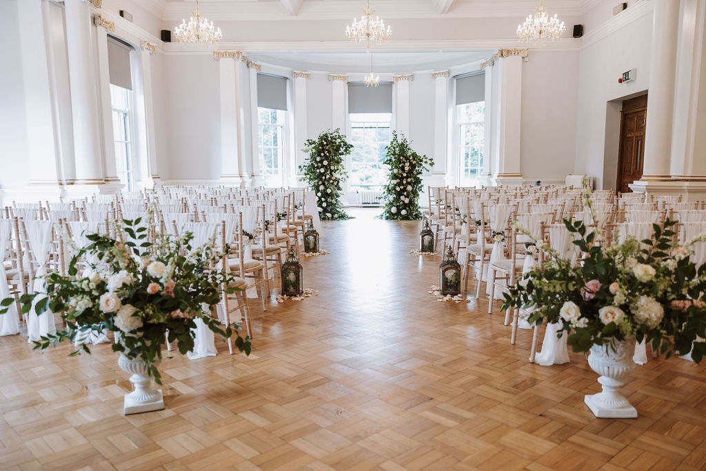 A bright, elegant wedding ceremony setup in a white hall with rows of white chairs, floral arrangements, and greenery at the front.