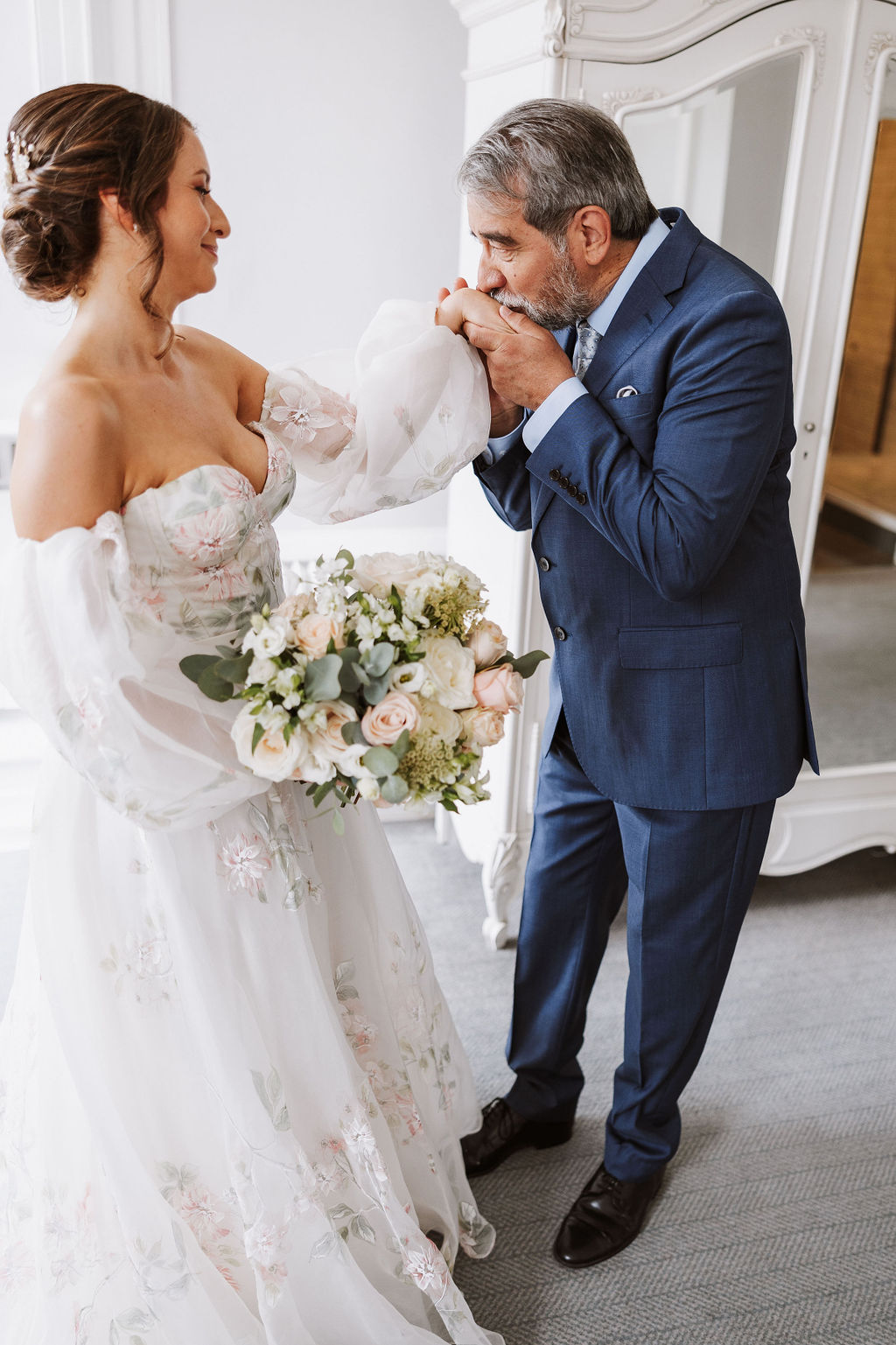 A man in a blue suit kisses the hand of a bride holding a bunch of flowers, both standing in a bright room with a white wardrobe in the background.