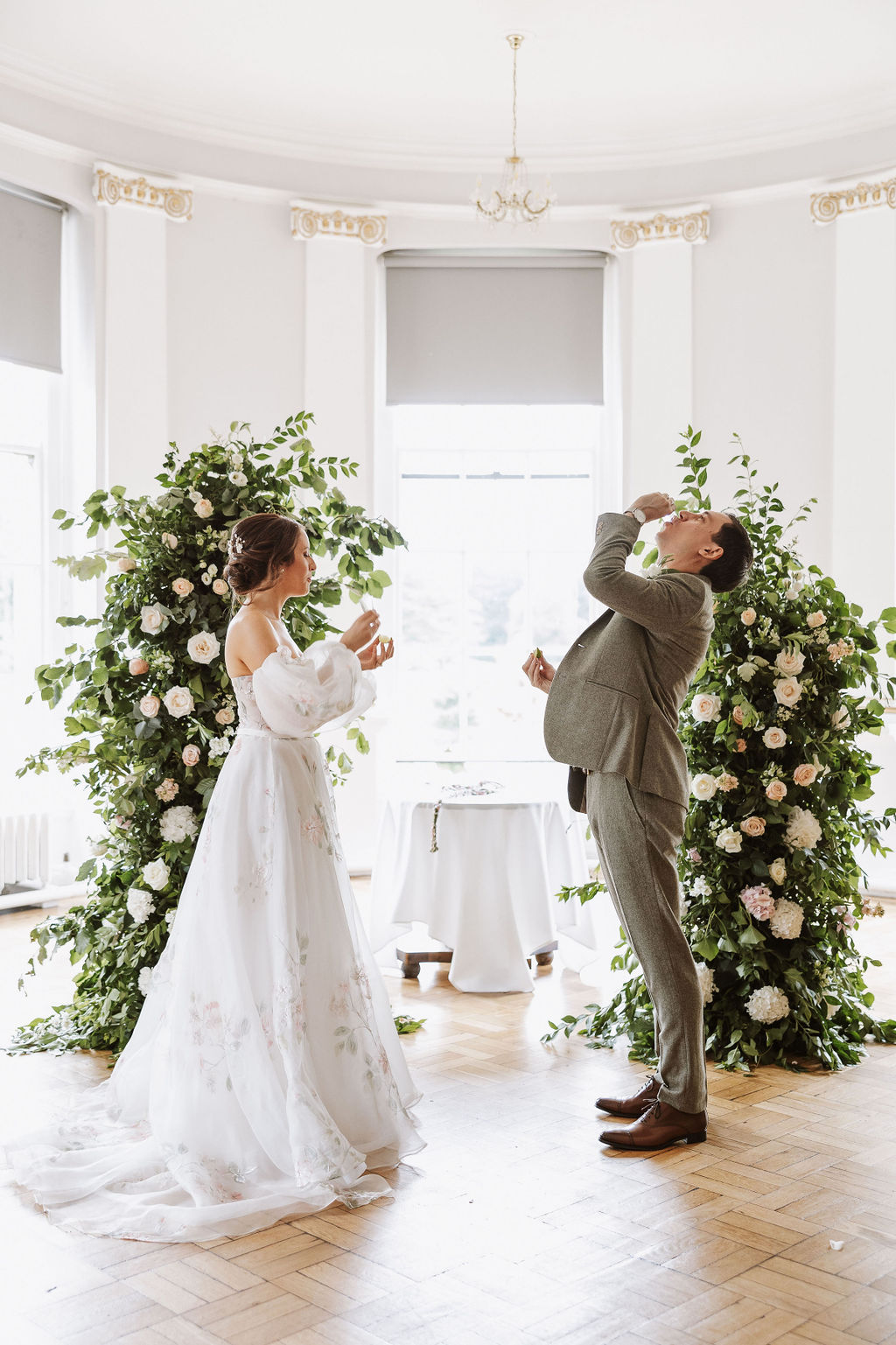 A bride and groom stand indoors in front of floral arrangements, both drinking from small glasses during a wedding ceremony.
