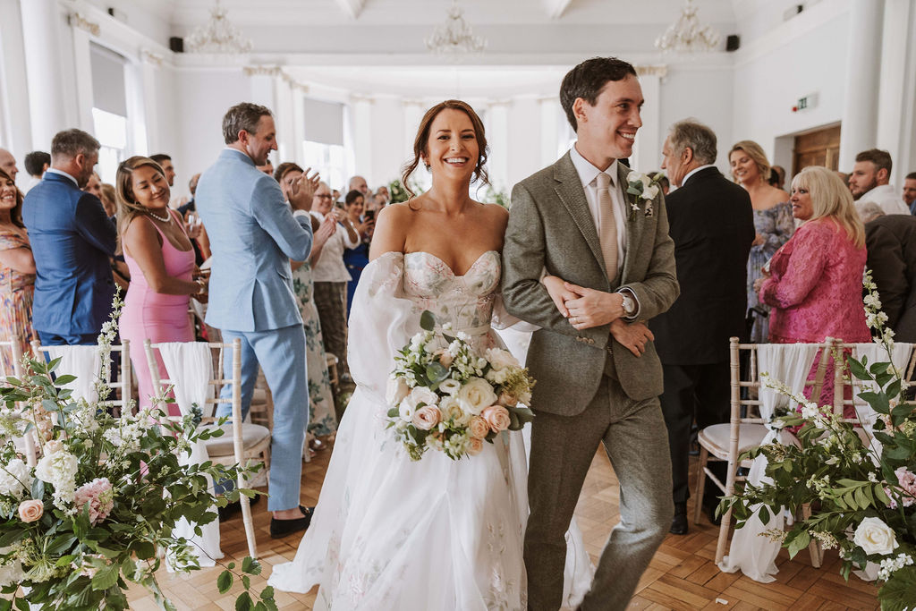 A bride and groom walk down the aisle smiling, surrounded by guests in a bright, elegantly decorated wedding venue with floral arrangements.
