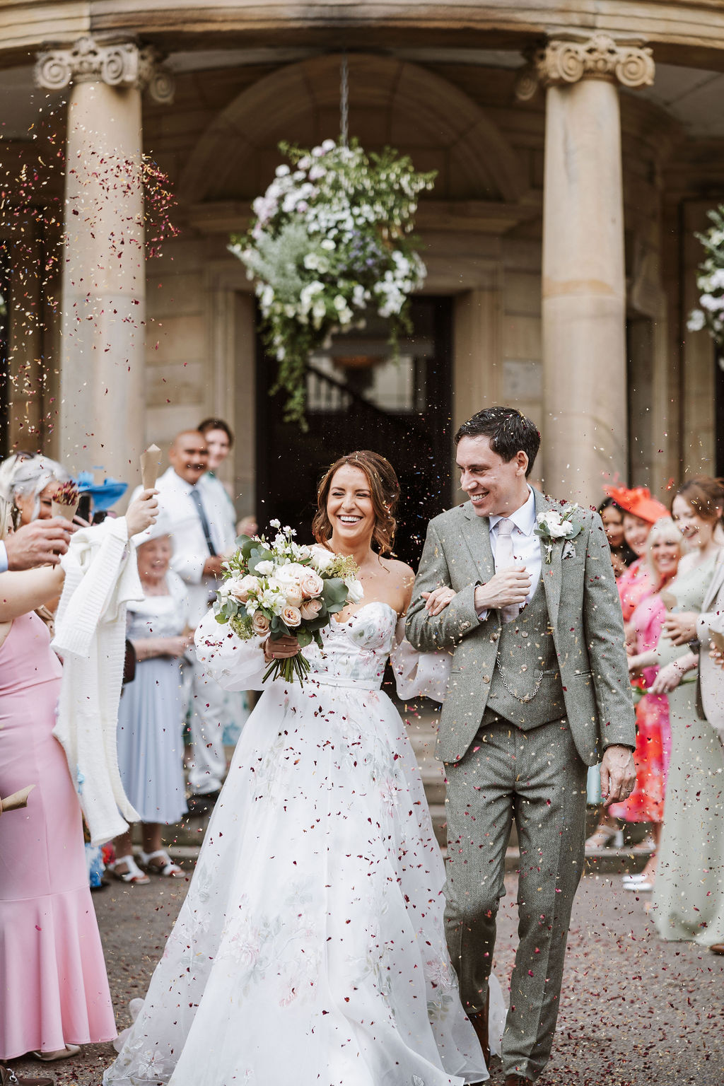 A bride and groom walk outdoors through a crowd of guests throwing confetti, smiling and holding hands, with a large floral arrangement in the background.
