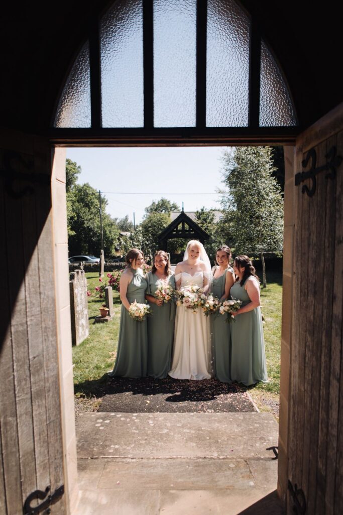 A bride in a white dress stands with four bridesmaids in green dresses outside a chapel, holding bouquets, viewed through an open doorway.