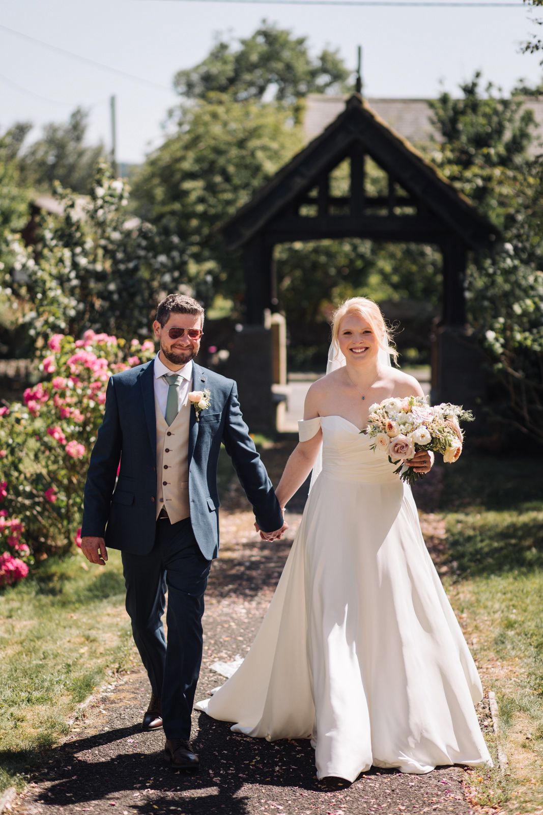 A bride in a white dress holds a bouquet and walks hand in hand with a groom in a blue suit outdoors on a sunny day at their romantic Healey Barn wedding.