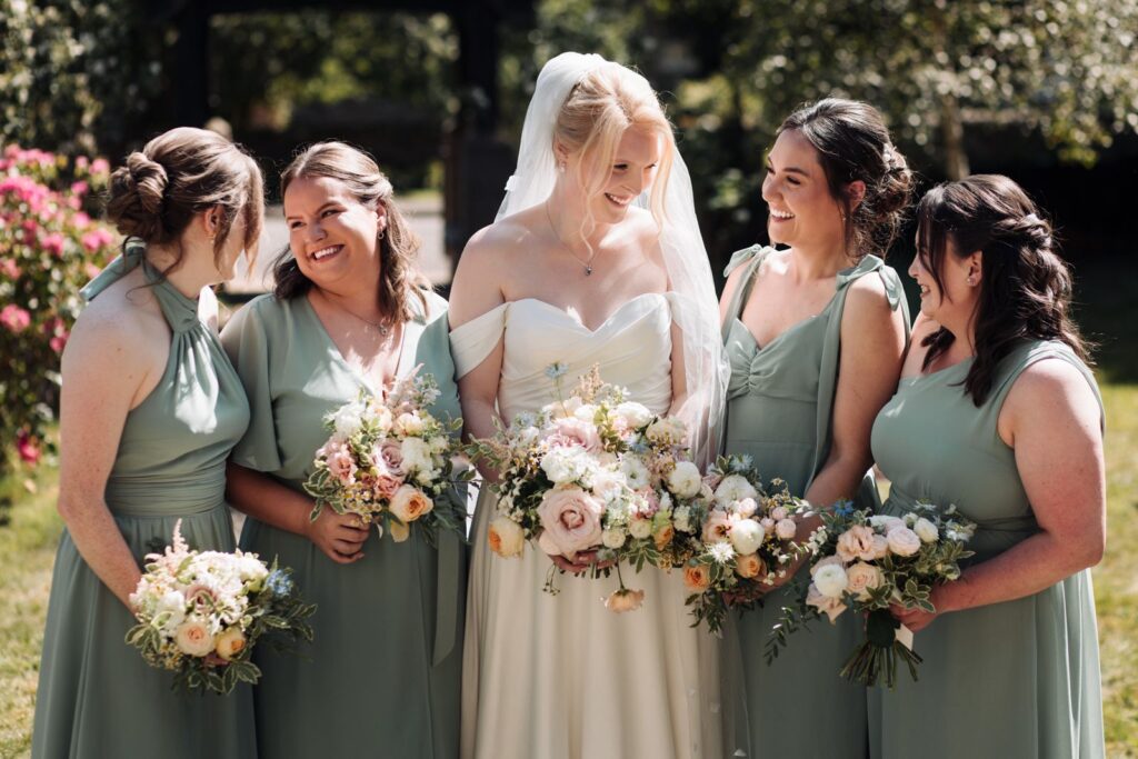 A bride in a white dress and veil stands outdoors in Northumberland with four bridesmaids in sage green dresses, all holding stunning bridal bouquets and wedding flowers, smiling warmly at each other.