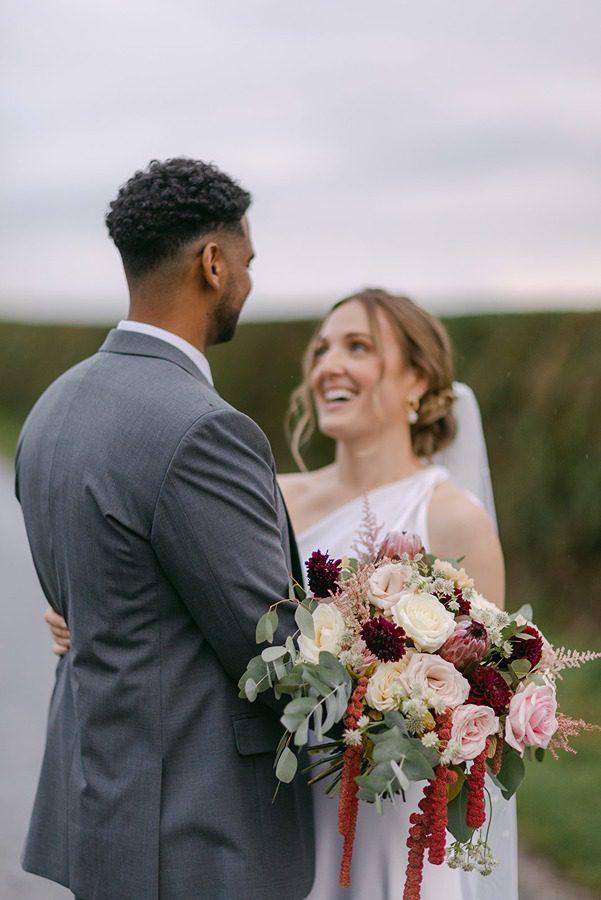 A bride holding a bouquet of mixed flowers smiles whilst standing close to a groom in a grey suit outdoors.