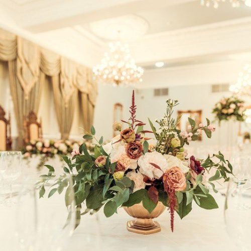 Elegant ballroom with chandeliers, ornate curtains, and a wedding flowers centrepiece on a round table set with glassware. Ornate chairs are visible in the background at Doxford Hall.