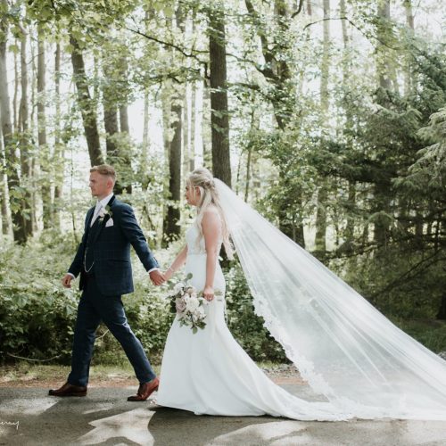 A bride and groom walk hand-in-hand along a wooded path, with the bride wearing a long veil and carrying a stunning bridal bouquet.