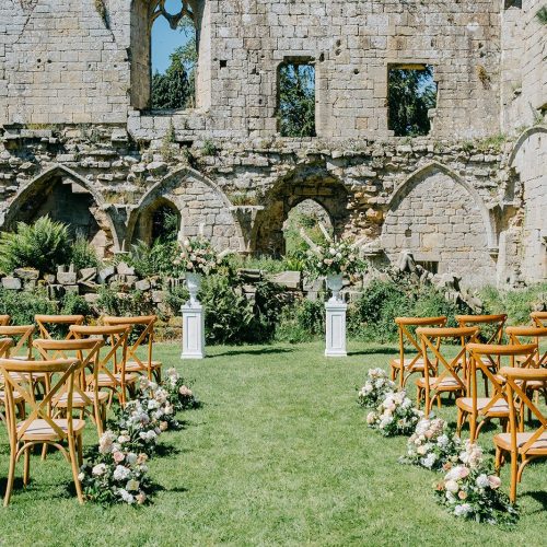 Rows of wooden chairs are arranged on grass with floral decorations, facing a small altar, set against the ancient stone ruins of Jervaulx Abbey—an enchanting outdoor wedding ceremony beneath open skies.