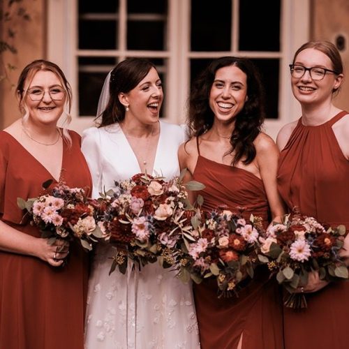 Four women pose in front of Middleton Lodge; one in a white wedding dress and the other three in matching reddish dresses, all holding floral bouquets and smiling after a beautiful Main House wedding ceremony.