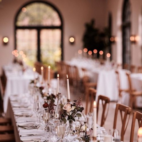 Long banquet tables at Middleton Lodge are set with white tablecloths, elegant glassware, exquisite wedding flowers, and lit taper candles in a beautifully decorated room with arched windows.