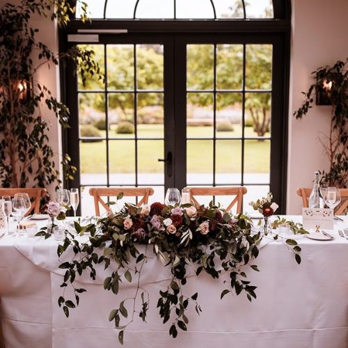 A long table set for a formal meal with a white tablecloth, glassware, and a floral centerpiece at a Middleton Lodge Fig House wedding reception, positioned in front of large windows overlooking a garden.
