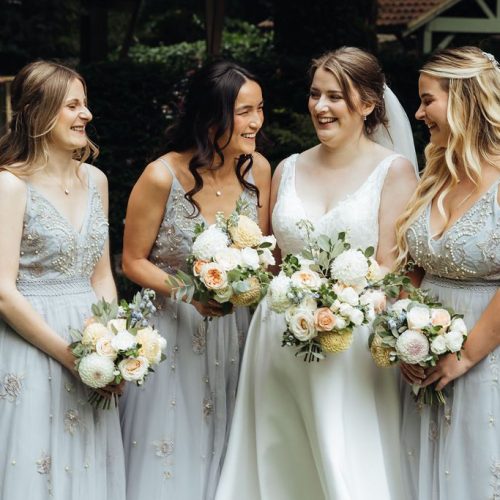 A bride in a white dress stands outdoors with three bridesmaids in light blue dresses, all holding beautiful Jesmond Dene House wedding flowers and smiling, each showcasing a stunning bridal bouquet.